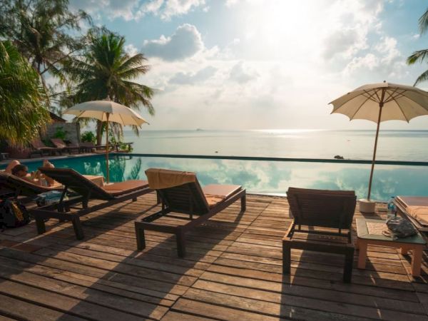 A serene poolside scene with lounge chairs, umbrellas, palm trees, and a tranquil ocean view under a partly cloudy sky.