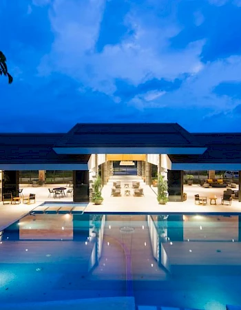 A luxurious building with a large swimming pool at night, surrounded by seating areas illuminated under a deep blue sky.
