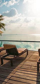 Sunlit poolside with lounge chairs, umbrellas, palm trees, and an infinity pool overlooking a calm ocean under a cloudy sky.