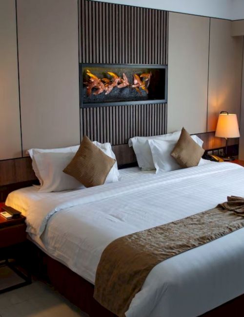 A neatly arranged hotel room with a large bed, pillows, lamps, a decorative towel arrangement, and a small fireplace on the wall.