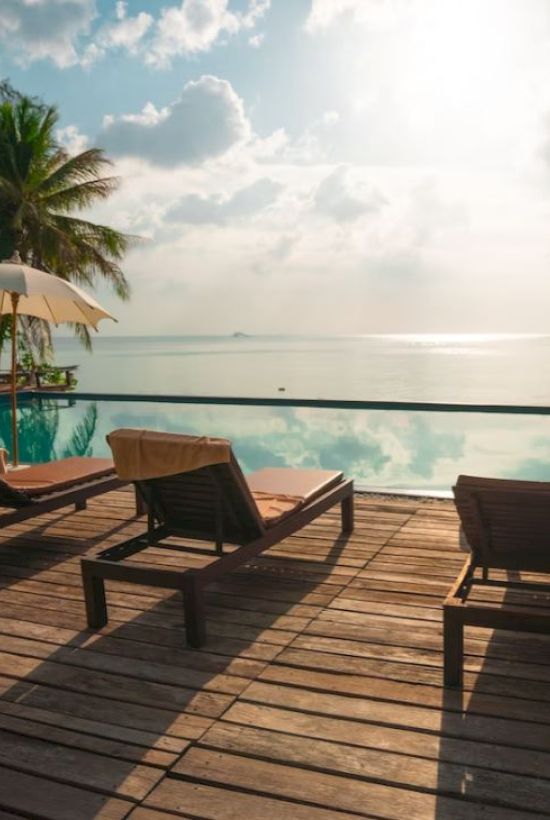 A serene poolside with lounge chairs and umbrellas overlooks a calm sea, surrounded by palm trees under a partly cloudy sky.