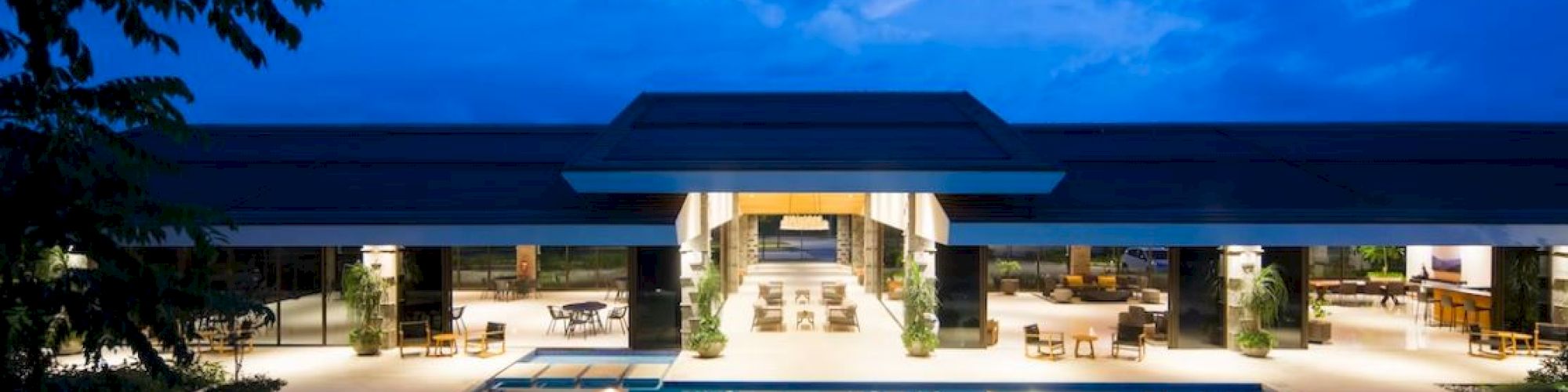 A luxurious outdoor pool area with a modern building, surrounded by trees and a clear evening sky, beautifully illuminated.