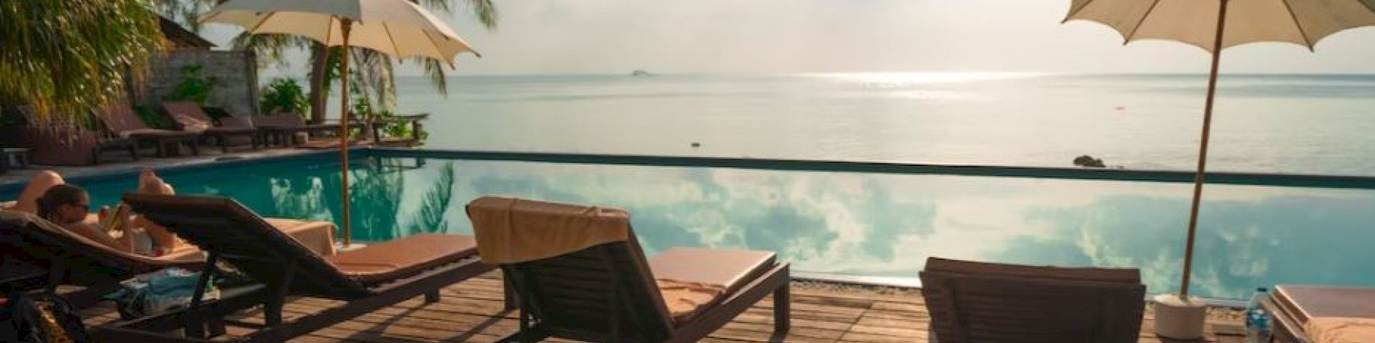 A serene infinity pool with lounge chairs and umbrellas, overlooking the ocean, surrounded by palm trees under a partly cloudy sky.
