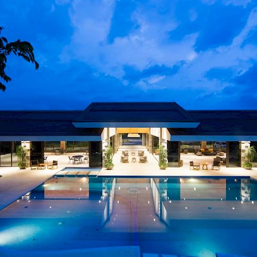 A luxurious, illuminated building with an inviting swimming pool, surrounded by trees under a vibrant evening sky.