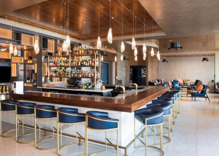 A modern bar with stylish seating, pendant lights, and a well-stocked shelf, set in a spacious, elegantly designed interior.