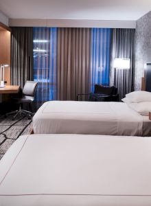 The image shows a modern hotel room with two beds, a desk, chair, and a wall-mounted TV, featuring stylish decor and lighting.