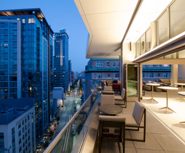 A modern urban balcony with seating, overlooking a city street lined with tall buildings, captured during twilight.