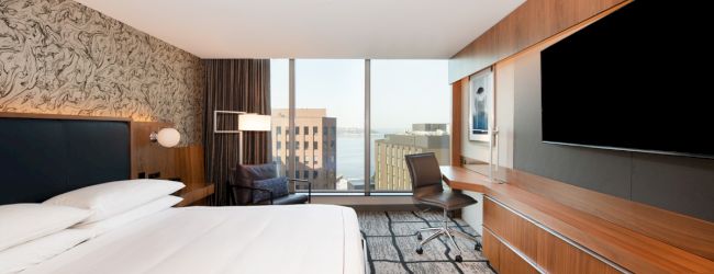 A modern hotel room with a bed, large TV, desk, chair, and a window view of buildings and water.