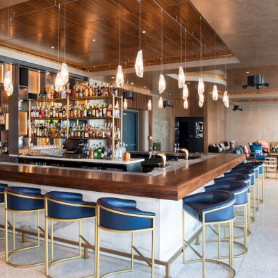 The image shows a modern bar with blue stools, a well-stocked liquor display, hanging lights, and a chic seating area in the background.