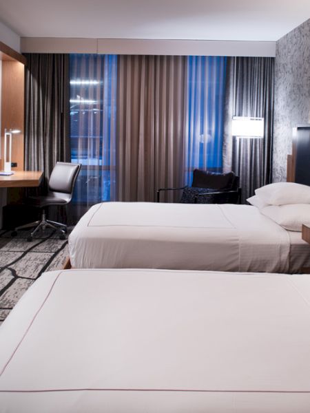The image shows a modern hotel room with two double beds, a large TV, a desk and chair by the window, and stylish decor.