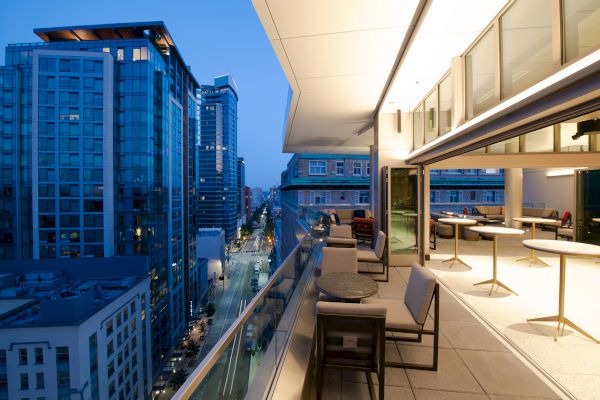 Rooftop terrace at dusk with modern furniture and cityscape views, showcasing tall buildings and illuminated street below.