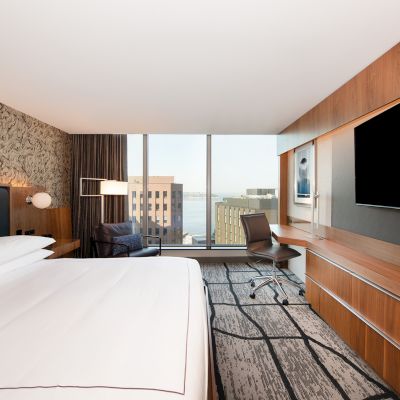 A modern hotel room with a large bed, flat-screen TV, desk, chair, and a view of buildings through the window.