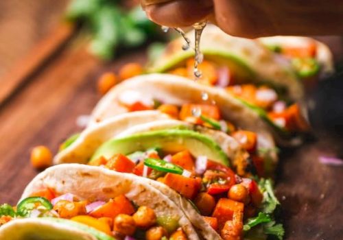 A hand squeezing lime over a row of tacos filled with chickpeas, avocado, and vegetables on a wooden board, garnished with cilantro.