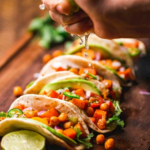A hand squeezing lime over a row of tacos filled with chickpeas, avocado, and vegetables on a wooden board, garnished with cilantro.