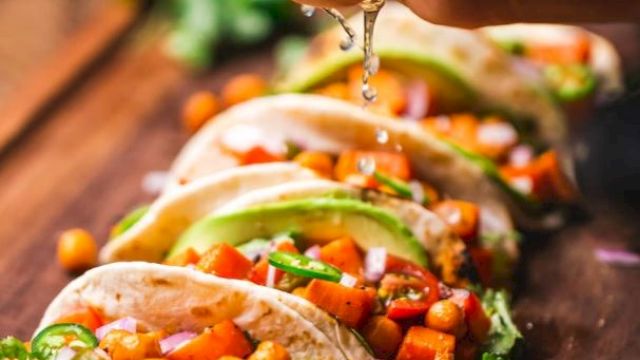 A hand squeezing lime over a row of tacos filled with chickpeas, avocado, and vegetables on a wooden board, garnished with cilantro.