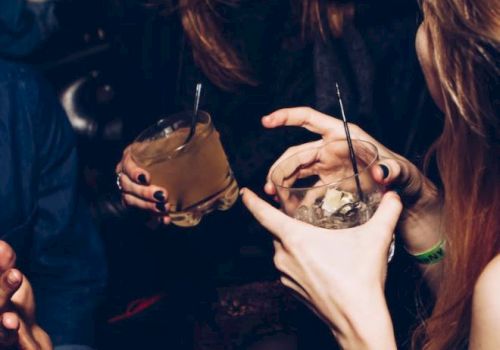 People are socializing at a party, holding drinks with straws, and laughing in a dimly lit setting.