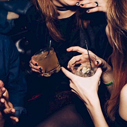People are socializing at a party, holding drinks with straws, and laughing in a dimly lit setting.