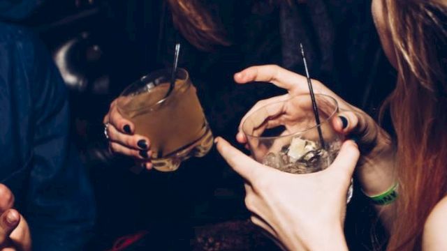 People are socializing at a party, holding drinks with straws, and laughing in a dimly lit setting.