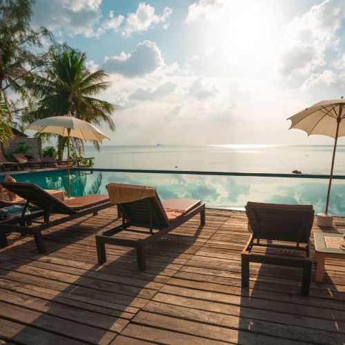 Lounge chairs and umbrellas by a pool overlooking the ocean, with palm trees and a sunny sky in the background.