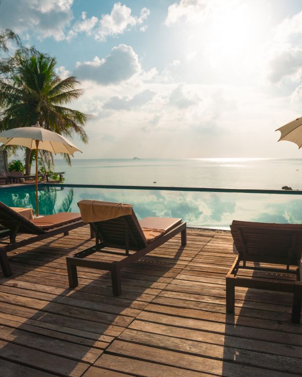 Lounge chairs and umbrellas by a pool overlooking the ocean, with palm trees and a sunny sky in the background.
