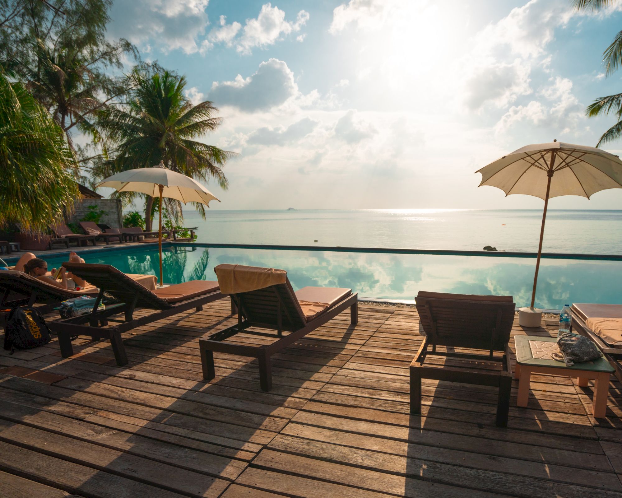 A beachside pool with lounge chairs and umbrellas, overlooking the ocean under a bright sky with scattered clouds.
