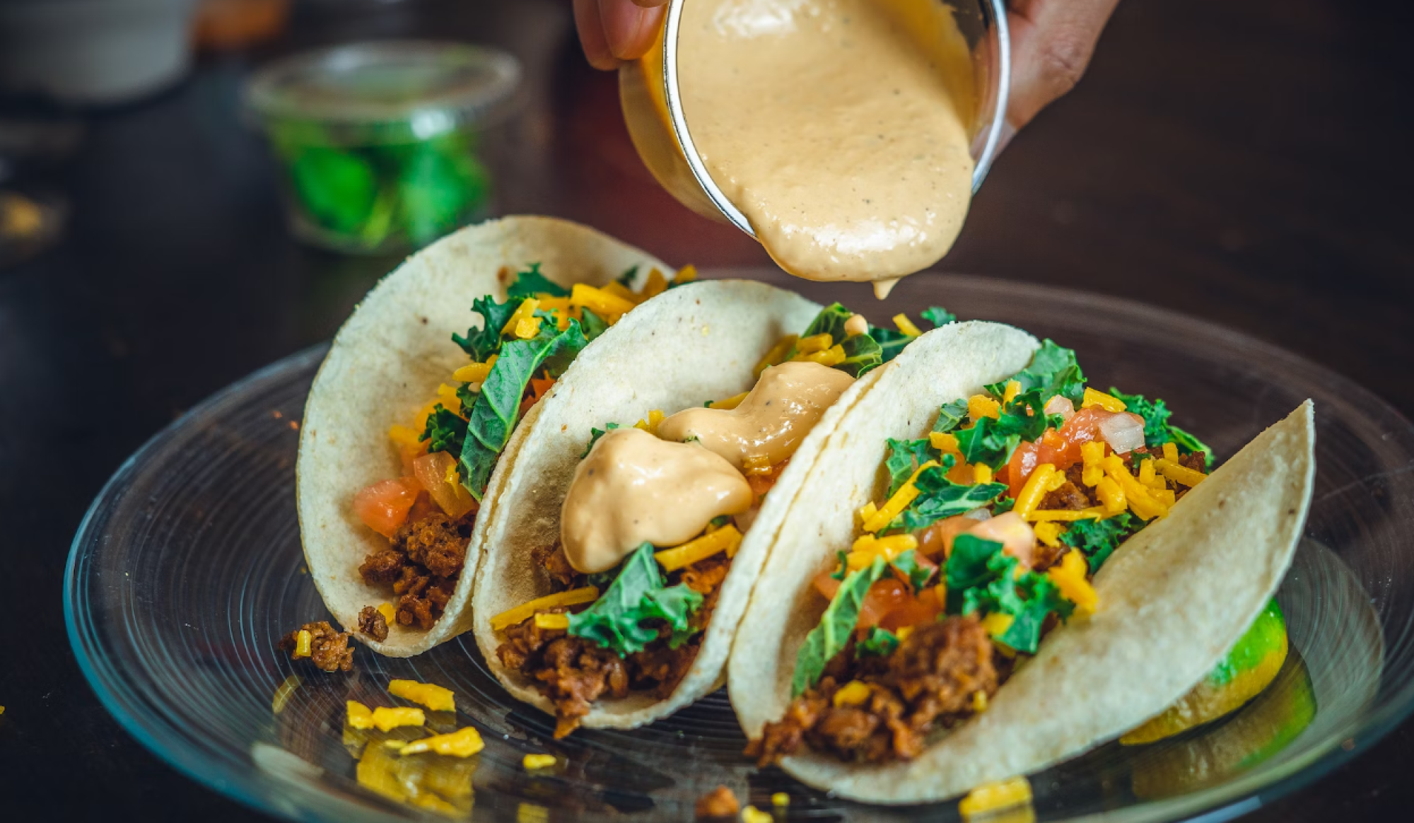 Three tacos with seasoned filling, topped with lettuce, cheese, and sauce being poured from a cup on a clear plate.