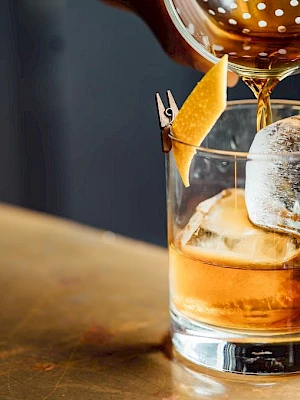 A whiskey cocktail being poured into a glass with ice and a lemon twist garnish, on a wooden surface.
