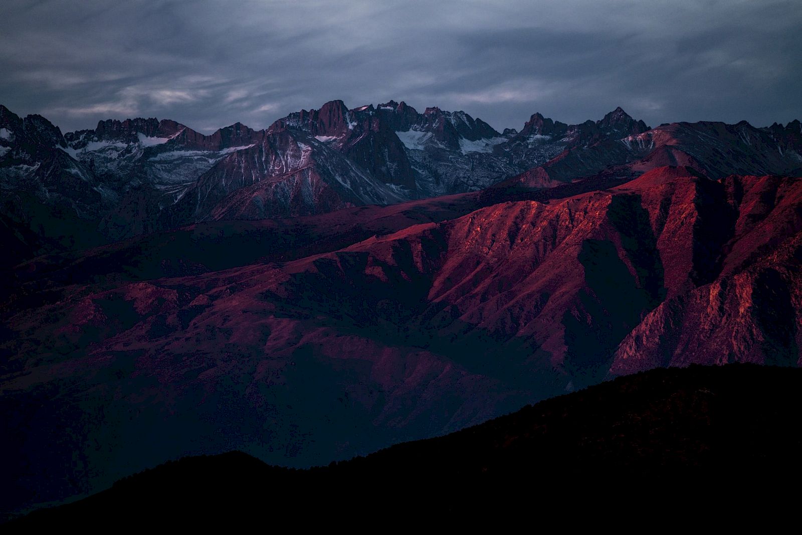 A mountain range with rocky peaks is bathed in red light under a cloudy sky.
