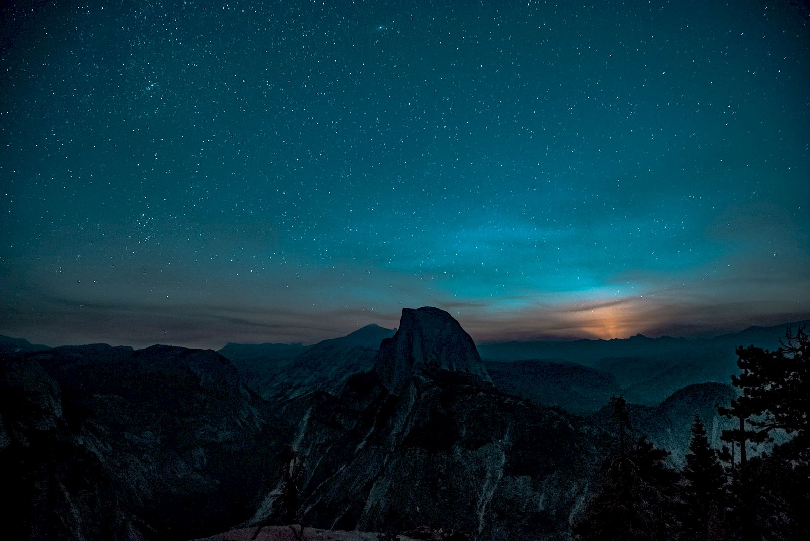 A night sky filled with stars over a mountain landscape, with a faint glow on the horizon, possibly from the rising or setting moon.