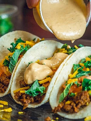 Three tacos filled with ground meat, shredded cheese, lettuce, and tomato, topped with sauce being poured over them.