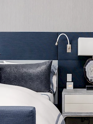 A modern bedroom with a blue headboard, white and blue pillows, a stylish nightstand with a decorative lamp, and a wall-mounted reading light.