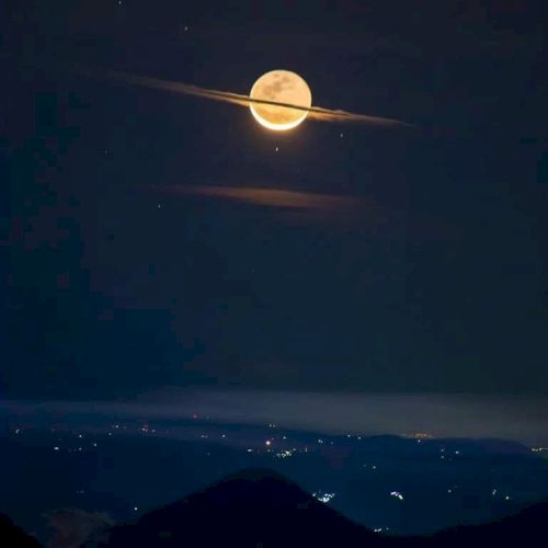A full moon appears with clouds resembling Saturn's rings above a silhouetted landscape with distant city lights.