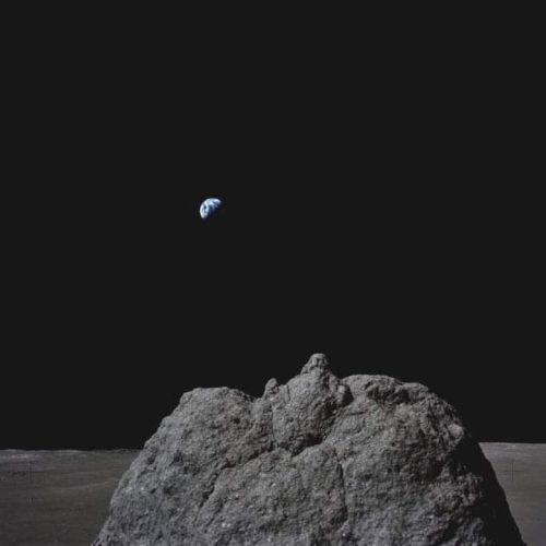 A large rock on a lunar surface with Earth visible in the distant black sky above.