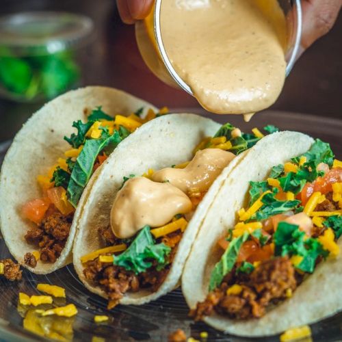 Three tacos filled with ground meat, cheese, tomatoes, and greens, topped with a creamy sauce being poured over them.