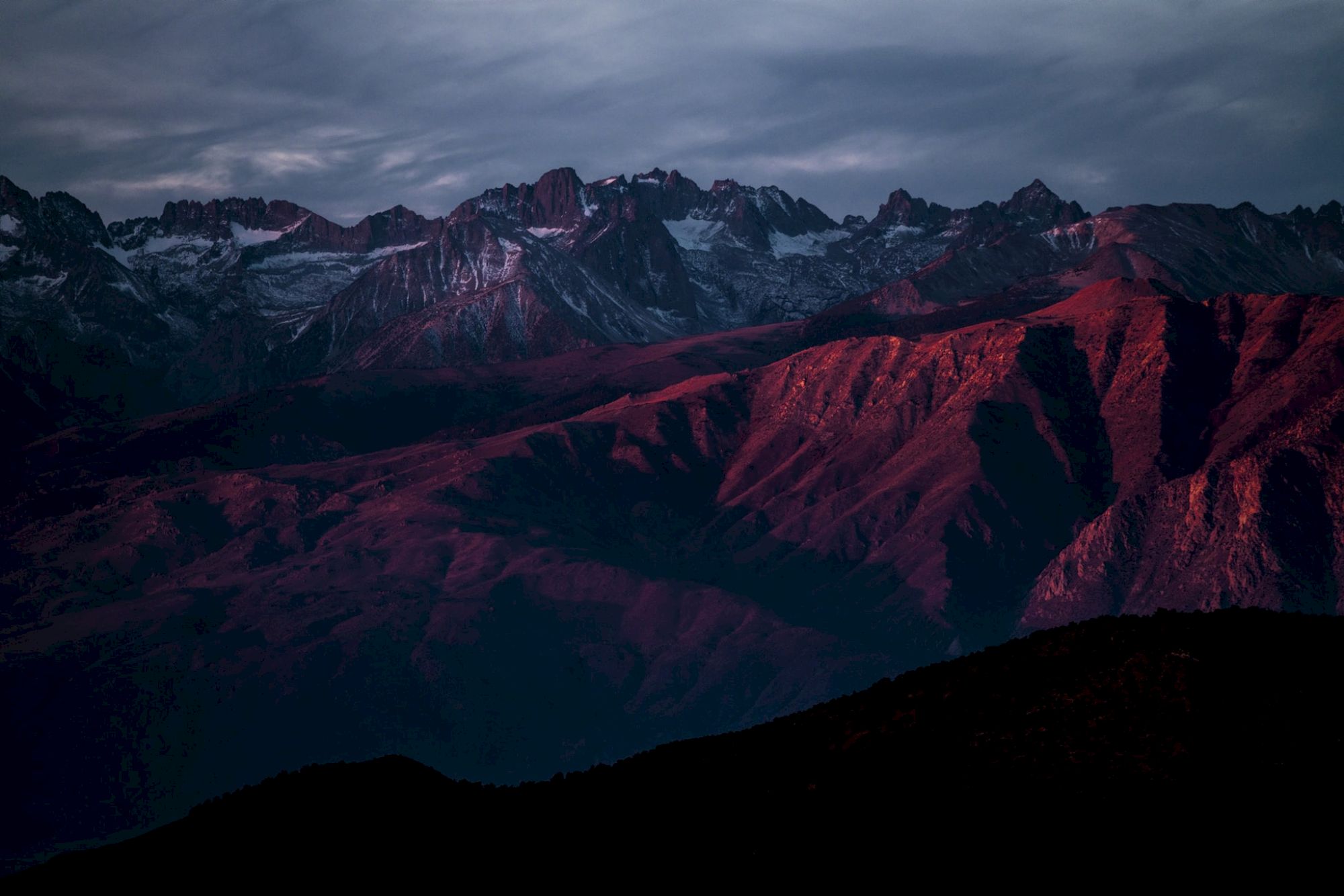 A mountain range with snow-capped peaks under a dramatic, cloudy sky at sunset, casting a red glow on the terrain.