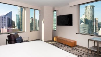 A modern hotel room with large windows offers a cityscape view, featuring a bed, TV, chair, and table, creating a cozy ambiance.