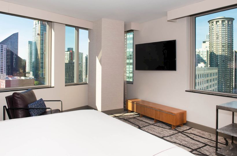 A modern hotel room with large windows offers a cityscape view, featuring a bed, TV, chair, and table, creating a cozy ambiance.