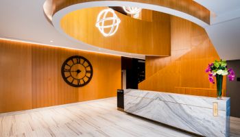 The image shows a modern lobby with a marble reception desk, wood paneling, and a large wall clock.