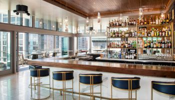 A modern bar with a wooden counter, stylish stools, and a well-stocked shelf of various drinks, in a bright setting.