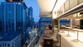 The image shows a modern balcony with tables and chairs overlooking a cityscape with high-rise buildings at dusk.