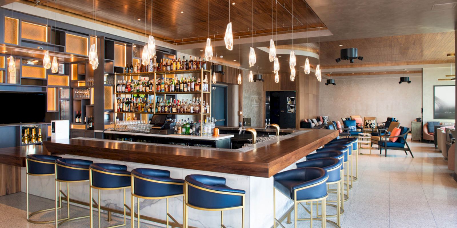 Stylish bar with modern design features, blue bar stools, hanging lights, and shelves filled with bottles; seating area in the background.