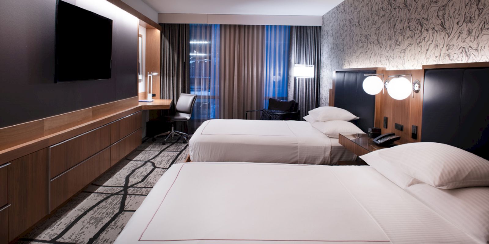 A modern hotel room with two beds, a TV, desk, and chair. Decor includes patterned walls and carpet, with soft lighting throughout.