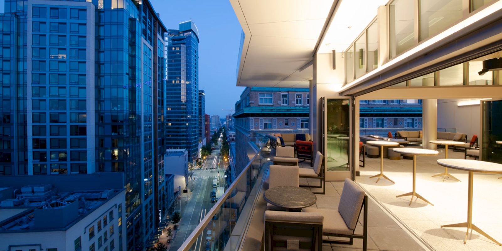 The image shows a modern balcony at dusk with tables, chairs, and city buildings in the background, viewed from a high vantage point.