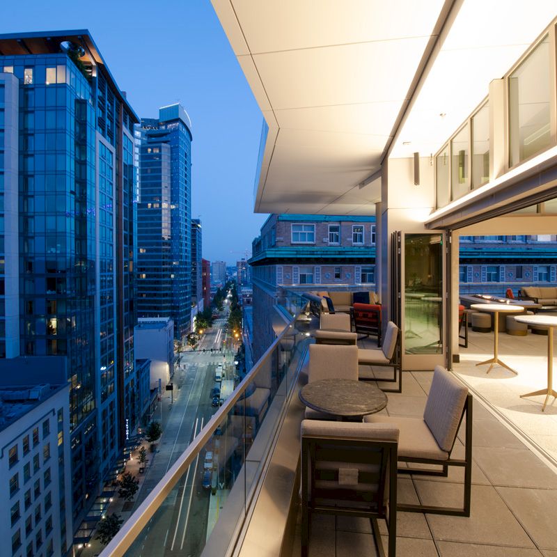 A modern cityscape at twilight with tall buildings; a sleek balcony with seating overlooks the street below, lit by streetlights.