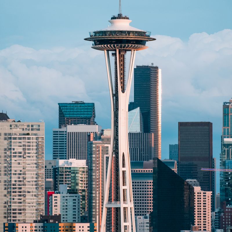 The image shows the Space Needle in Seattle, surrounded by modern skyscrapers under a blue sky and clouds in the background.