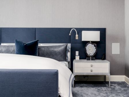 A modern bedroom features a navy upholstered bed with white bedding, a bedside table with a lamp and decor piece, on a plush grey carpet.