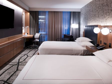 A hotel room with two beds, wall-mounted TV, desk, chair, lamps, and patterned wall.