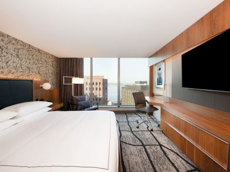 A modern hotel room with a large bed, desk, chair, TV, and a view of buildings and water outside the window, decorated in neutral tones.