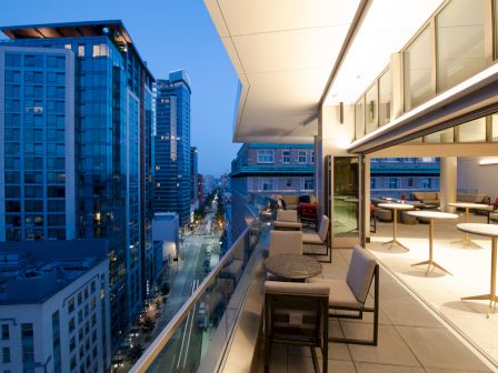 A modern rooftop terrace overlooks a cityscape at dusk, featuring outdoor seating and open-air dining with illuminated buildings in view.