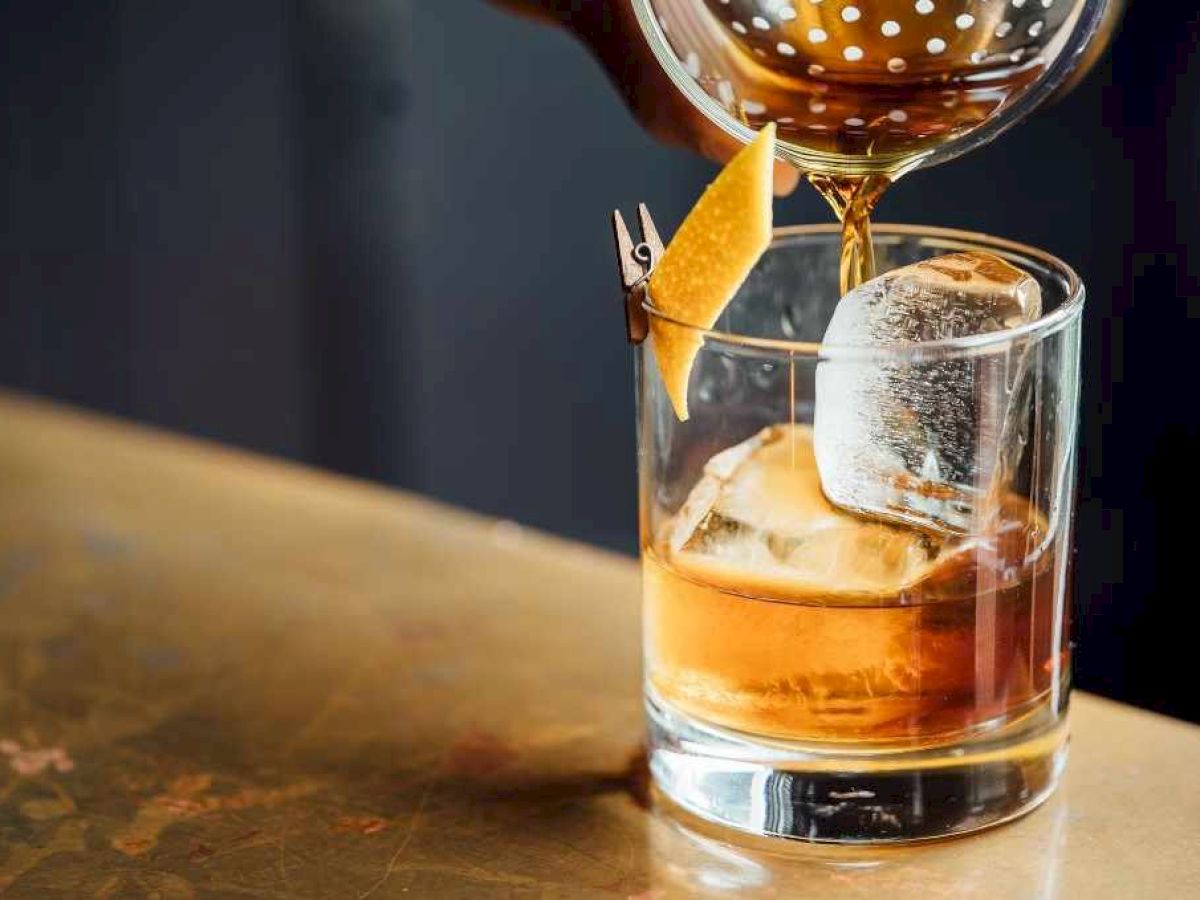 A cocktail is being poured into a glass with ice and a citrus peel garnish, set on a bar counter.